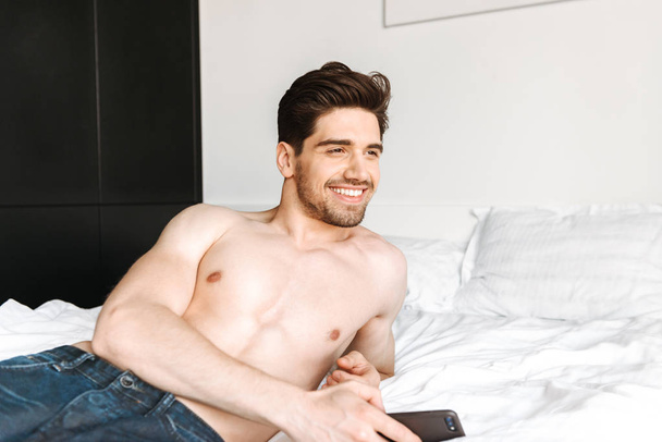 Smiling shirtless man holding mobile phone while laying on bed at home - Zdjęcie, obraz