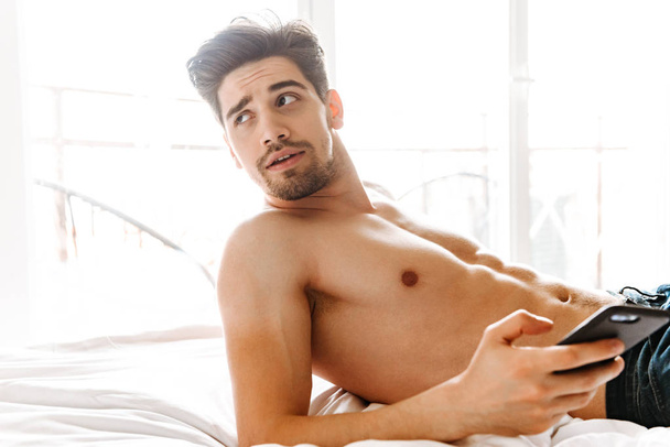Charming shirtless man holding mobile phone while laying on bed at home - Фото, зображення