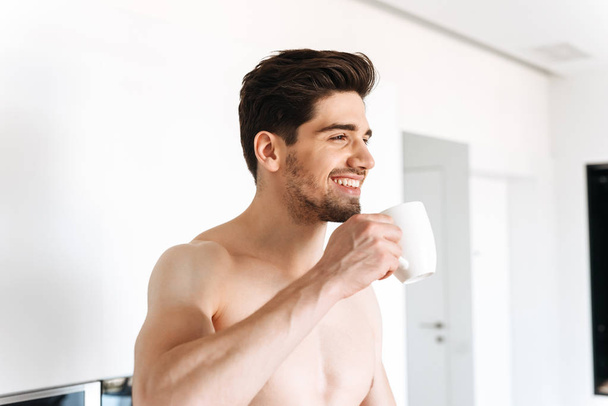 Happy shirtless man drinking coffee while standing at the kitchen - Photo, image