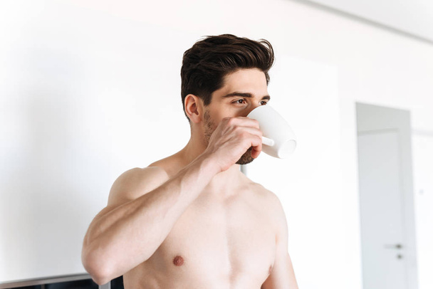 Handsome shirtless man drinking coffee while standing at the kitchen - Fotografie, Obrázek