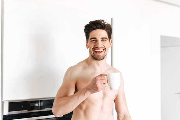 Excited shirtless man drinking coffee while standing at the kitchen - Fotografie, Obrázek