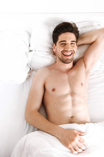 Photo of young handsome cheerful naked man lies in bed indoors at home looking camera. - Foto, Bild