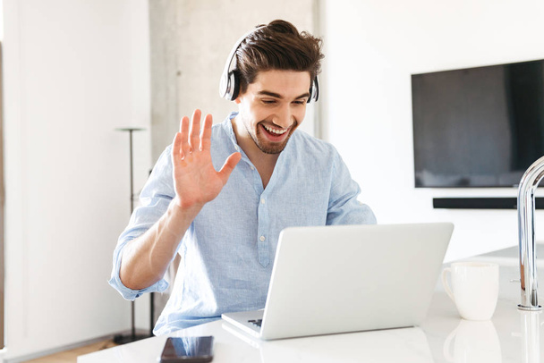 Portrait of a smiling young man in headphones waving at laptop computer while sitting at home - Photo, Image