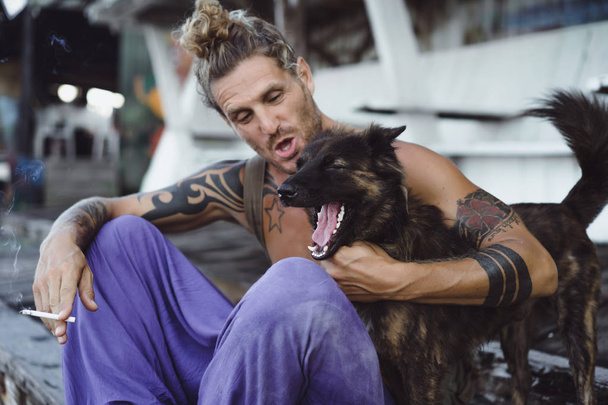 Handsome adult man with long blond hair and tattoos playing with dog - Фото, изображение