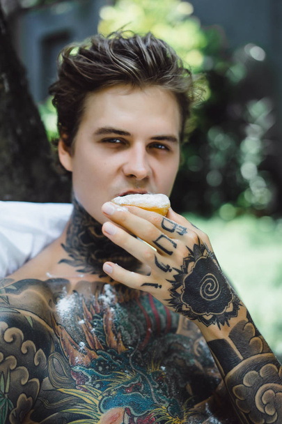 young handsome tattooed man having breakfast in bed in the open air outdoors in the garden - Fotó, kép