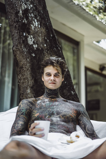 young handsome tattooed man having breakfast in bed in the open air outdoors in the garden - Photo, image