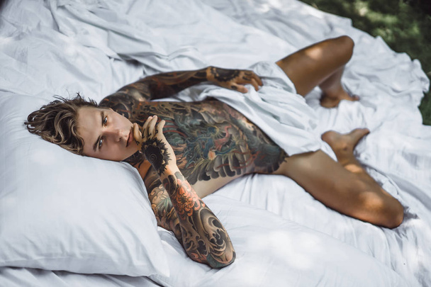 Handsome young tattooed man relaxing on white bed - Photo, image