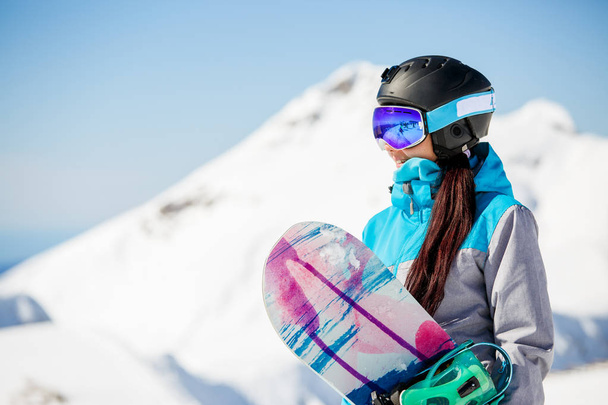 Photo of young woman tourist in helmet looking away with snowboard in hands against background of snow mountains - 写真・画像