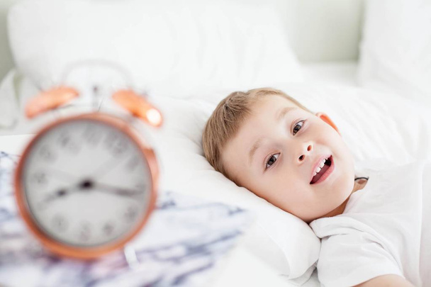目覚まし時計は、ベッド、ベッドサイドのテーブルの上です。小さな男の子は白いベッドの上で横たわっています。起きてください。朝。居心地の良い。学校. - 写真・画像
