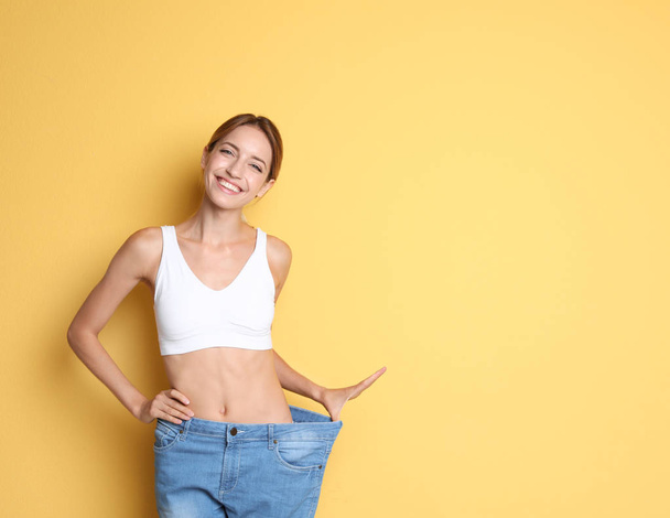 Young slim woman in old big jeans showing her diet results on color background - Φωτογραφία, εικόνα
