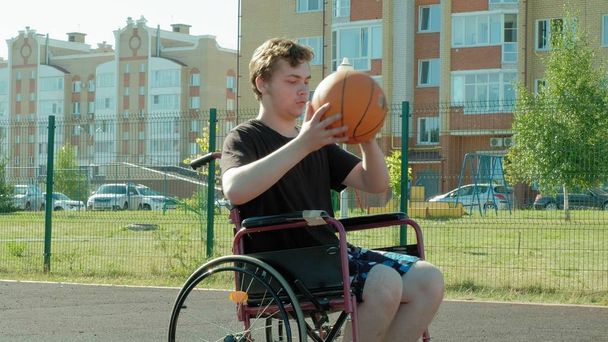 Homem com deficiência joga basquete de sua cadeira de rodas, ao ar livre
 - Foto, Imagem