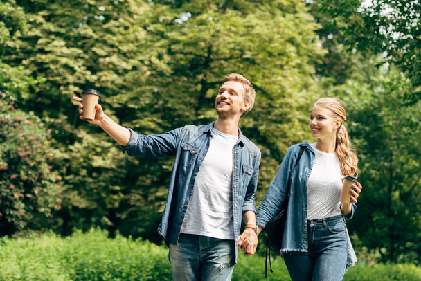 Szczęśliwa Młoda para z papierowych kubków kawy spaceru przez park razem i wskazując gdzieś - Zdjęcie, obraz