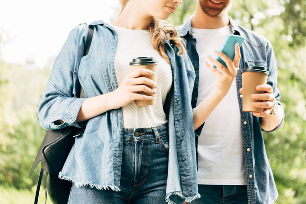 plan recadré de jeune couple avec des tasses en papier en utilisant smartphone ensemble au parc
 - Photo, image