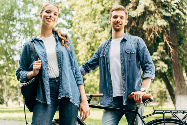 happy young couple with vintage bicycle standing at park and looking at camera - Фото, зображення