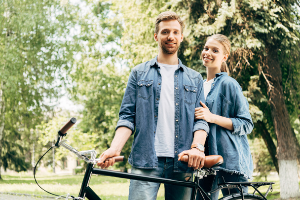 beautiful young couple with vintage bicycle standing at park and looking at camera - Foto, Imagem