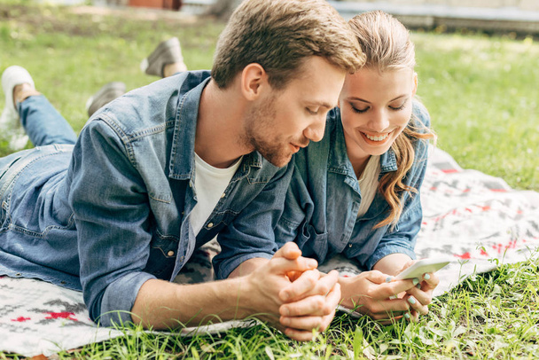 smiling young couple lying on grass at park and using smartphone - Foto, imagen