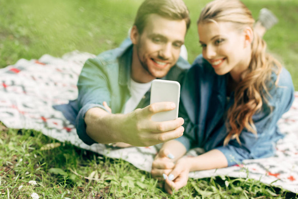 beautiful young couple lying on grass at park and taking selfie - Photo, Image