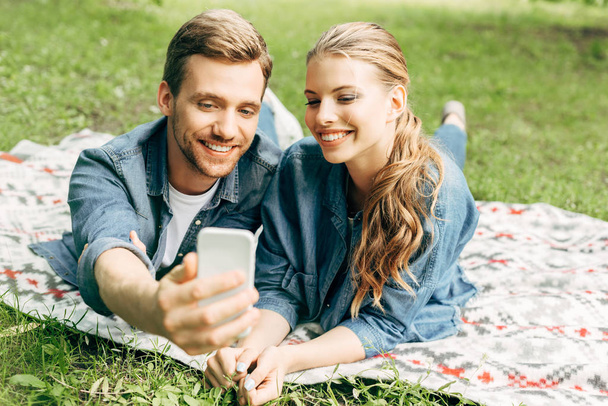 smiling young couple lying on grass at park and taking selfie - Photo, Image
