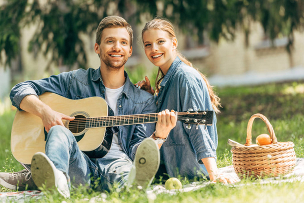 jovem feliz tocando guitarra para sua namorada sorridente durante piquenique no parque e olhando para a câmera
 - Foto, Imagem