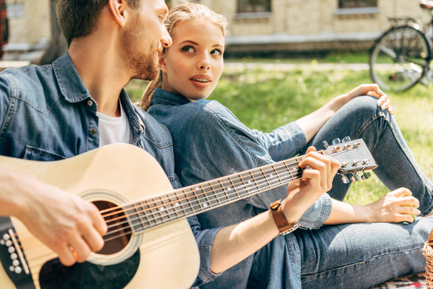 handsome young man playing guitar for his girlfriend while relaxing at park - Foto, Imagen