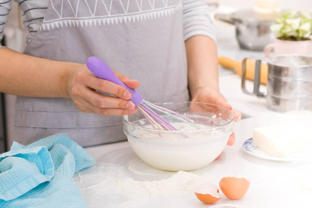Frau in Schürze mixt Eier in Keramikschüssel für Teig auf Holztisch in der Küche. Kochprozess für schrittweises Rezept. - Foto, Bild