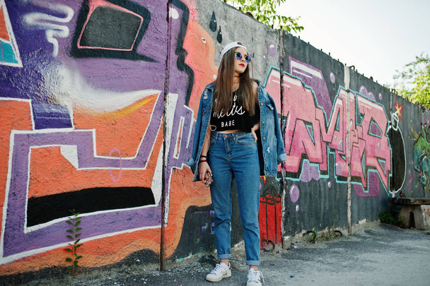 Stylish casual hipster girl in cap, sunglasses and jeans wear, listening music from headphones of mobile phone against large graffiti wall. - 写真・画像