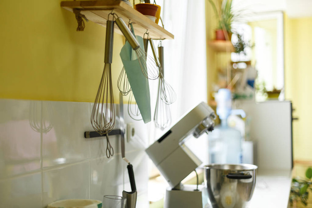 Beater whisk, confectioner's bag and other kitchen accessories hang on yellow kitchen wall. - Фото, изображение