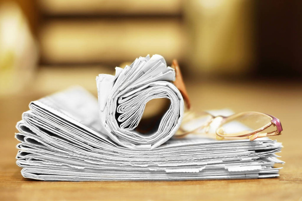 Складные и свернутые газеты сложены в кучу и очки для чтения на деревянном столе, вид сбоку
  - Фото, изображение