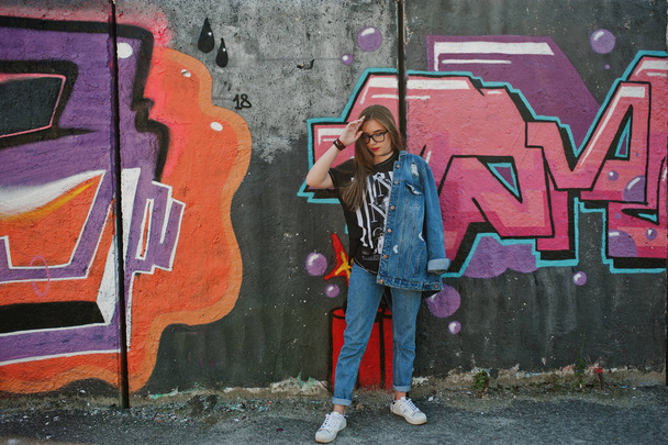 Stylish casual hipster girl in jeans wear and glasses against large graffiti wall. - Photo, Image
