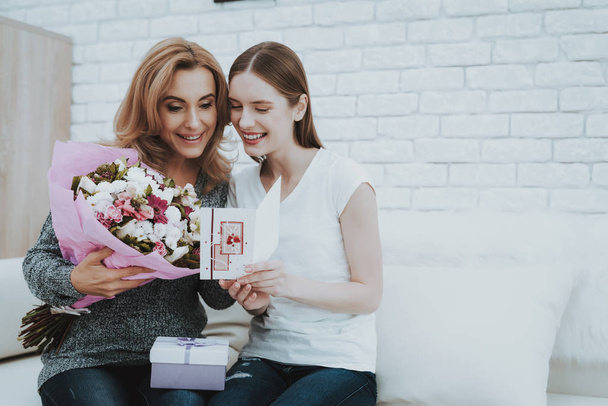 Smiling Daughter Gives Mother Flowers and Gift. Relationship in Family. Holiday at Home. Happy Mother. Friendly Relations. Celebrating Together. Happiness in Family Concept. Presenting Gift. - Zdjęcie, obraz