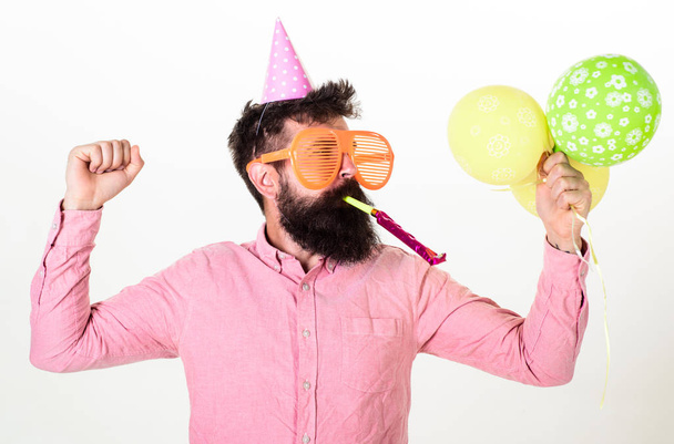 Guy in party hat with air balloons celebrates. Man with beard and mustache on busy face blows into party horn, white background. Hipster in giant sunglasses celebrating birthday. Celebration concept - Foto, immagini