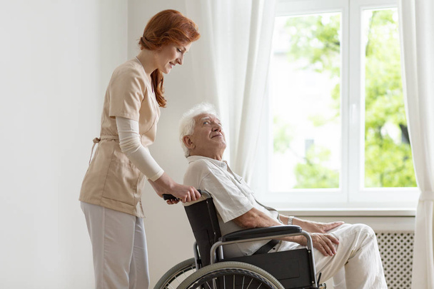 Smiling nurse supporting disabled senior man in the wheelchair - Valokuva, kuva