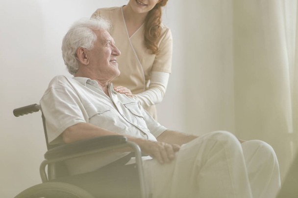 Low angle on smiling and happy disabled elderly man in a wheelchair and his caregiver in a uniform - Foto, immagini
