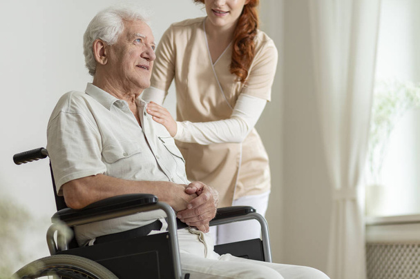 Caregiver supporting sick elderly man in the wheelchair during stay in the hospice - Fotó, kép