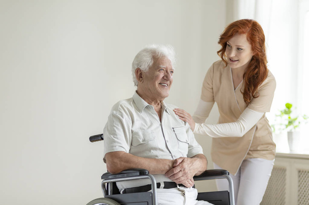 Friendly nurse supporting smiling paralyzed senior man in a wheelchair - Фото, изображение