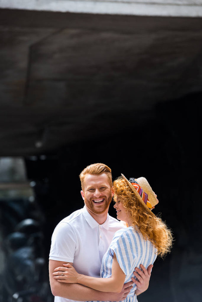 smiling redhead man embracing girlfriend in straw hat at city street - Foto, imagen