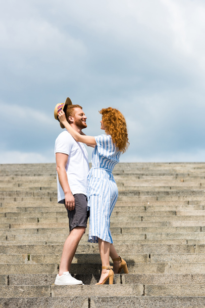 attractive redhead woman putting on own straw hat on boyfriends head on stairs  - Photo, Image