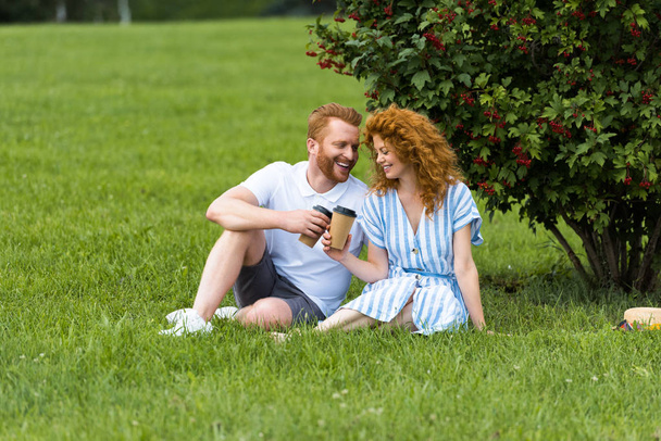 happy redhead couple clinking by paper cups with coffee on grass in park - Фото, изображение