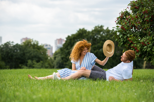 笑顔赤毛の女性が公園の芝生の上の彼氏の頭の上の自身の麦わら帽子に置くことにしようとしています。  - 写真・画像