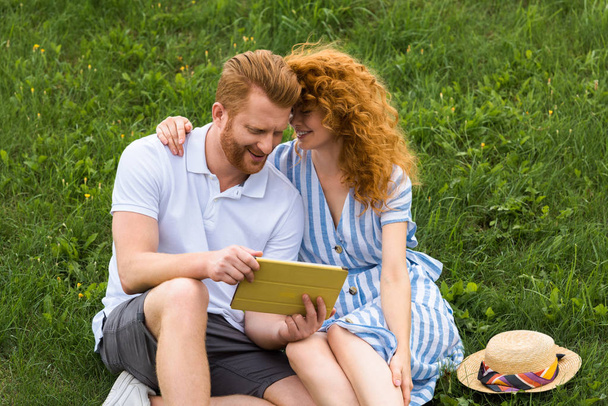happy redhead couple with smartphone sitting on grassy meadow - Photo, Image