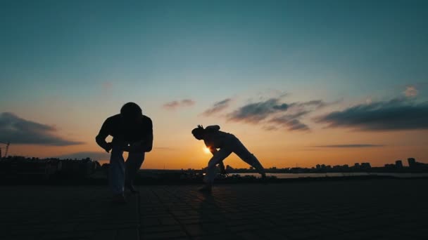Két sportos férfi, aki táncol a brazil capoeira, a háttérben a naplemente nyári sziluettek - Felvétel, videó