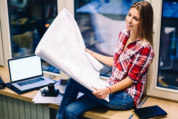 Smiling woman holding papers with drawings satisfied with completing blueprint before deadline sitting on window sill near laptop computer with mock up screen, female architect holding sketch - Photo, Image