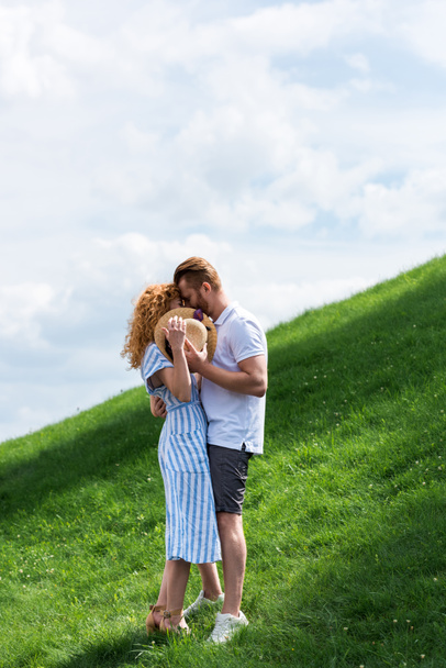 young redhead woman covering face by straw hat while kissing with boyfriend on green hill  - Foto, imagen