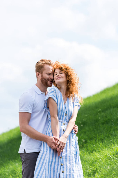 happy redhead man embracing girlfriend from behind on grassy hill  - Fotoğraf, Görsel