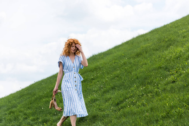 attractive redhead woman carrying shoes on grassy hill  - Фото, зображення