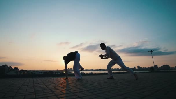 Männer mit sportlichem Körperbau widmen sich der brasilianischen Kampfkunst Capoeira im Freien, auf dem Asphalt, vor dem Hintergrund der Schönheit der Promenade und des sommerlichen Sonnenuntergangs - Filmmaterial, Video