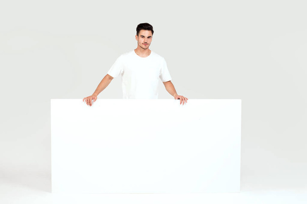Young handsome guy holding blank white banner, signboard, isolated on white studio background. - Photo, Image