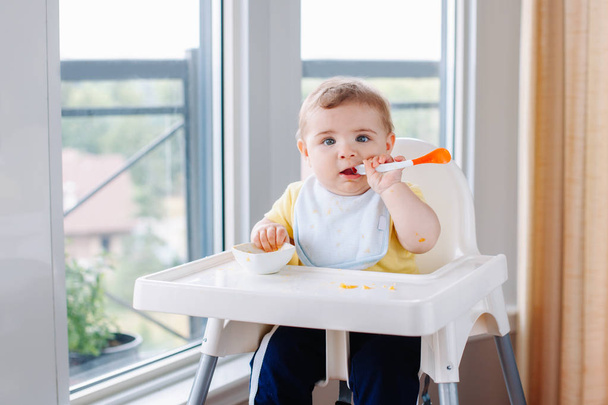 Portrait of cute adorable Caucasian child boy with dirty messy face sitting in high chair eating apple puree with spoon. Everyday home childhood lifestyle. Infant trying supplementary baby food - Φωτογραφία, εικόνα