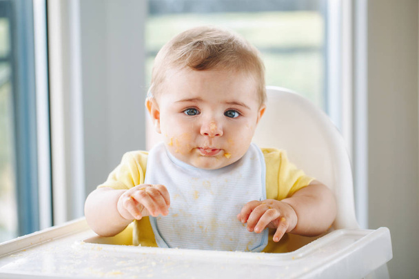 Portrait of cute adorable Caucasian child boy with dirty messy face sitting in high chair eating apple puree with fingers. Everyday home childhood lifestyle. Infant trying supplementary baby food - Photo, Image