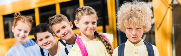 Panoramaaufnahme einer Gruppe entzückender Schüler, die in die Kamera schauen, während sie vor dem Schulbus in der Reihe stehen - Foto, Bild